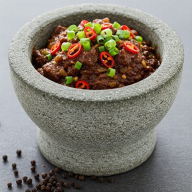 Grijze stenen kom met pittige saus, groene uien en rode pepers bovenop.